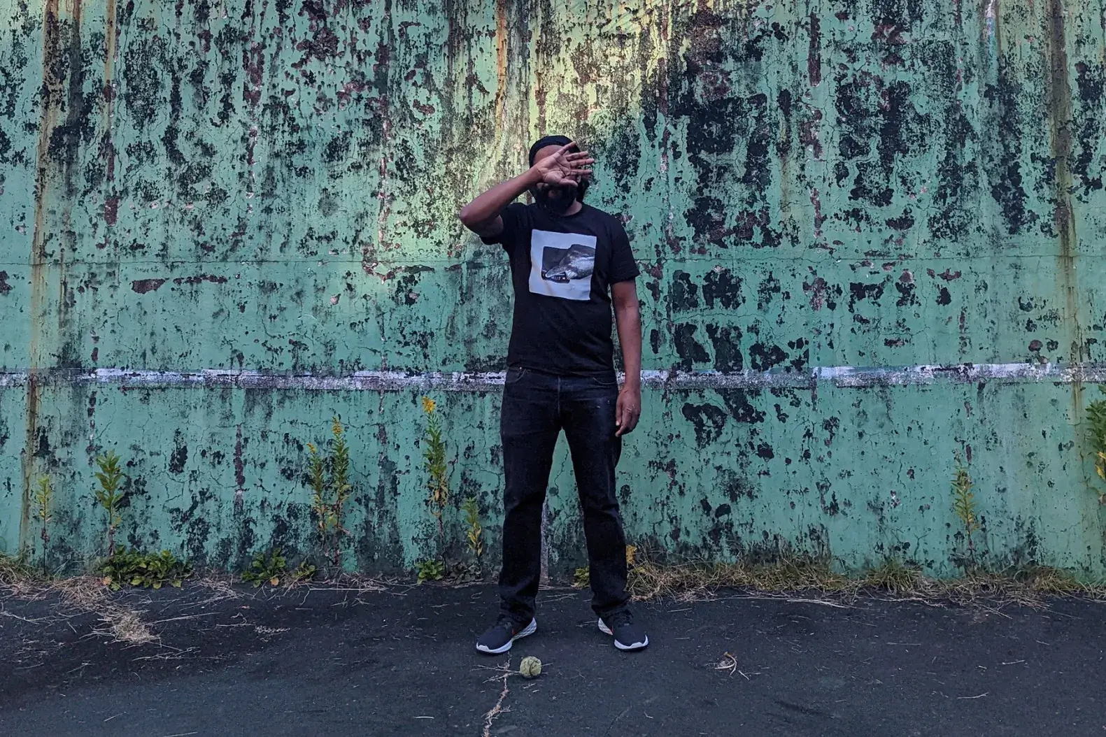 Artist Billy Woods covering their face in front of a green and black wall. 