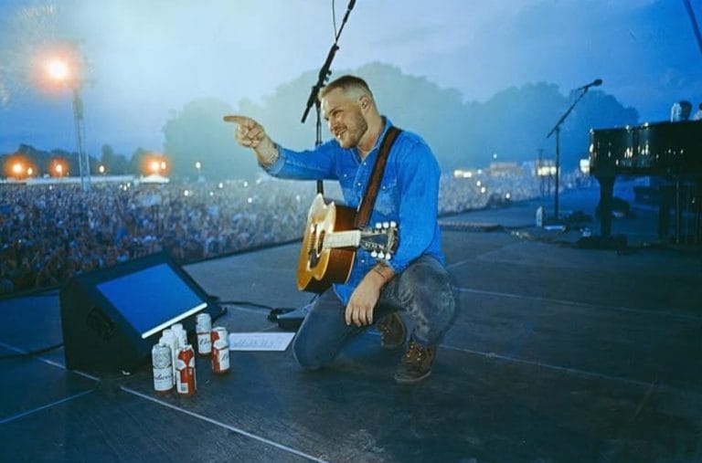 Country singer Zach Bryan kneels on stage with a guitar, pointing toward the crowd during an outdoor concert at dusk, with a large audience and stage equipment visible in the background.