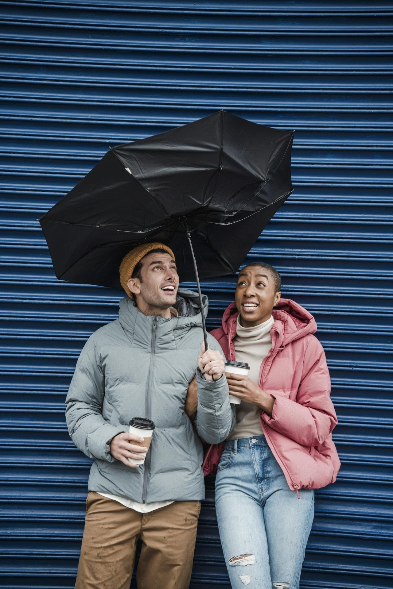 Diverse excited man and woman in outer weather under umbrella caught in gust of wind near blue wall on street of sf