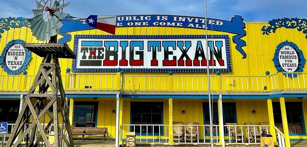 Exterior of The Big Texan Steak Ranch with a large sign on a bright yellow building, wooden porch seating, and a windmill display out front.