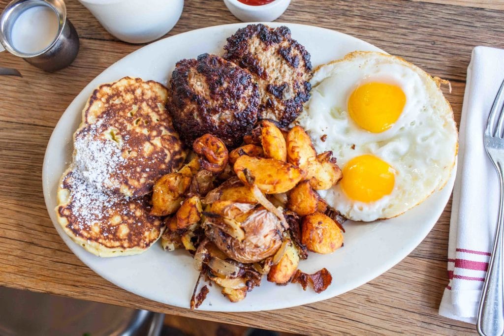 San Francisco Restaurants brunch plate with pancakes, eggs, breakfast potatoes, and grilled sausage patties on a wooden table.