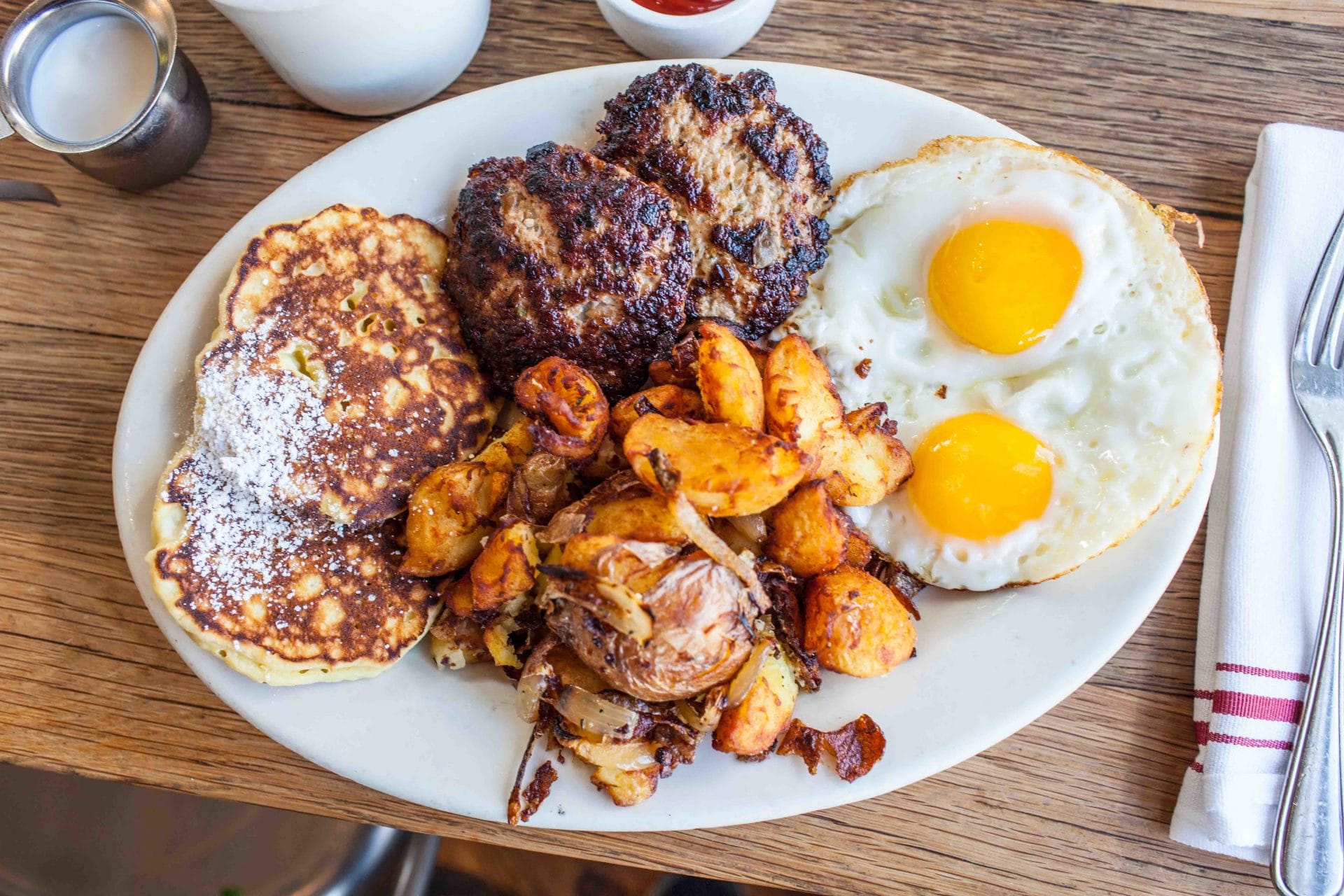 San Francisco Restaurants brunch plate with pancakes, eggs, breakfast potatoes, and grilled sausage patties on a wooden table.
