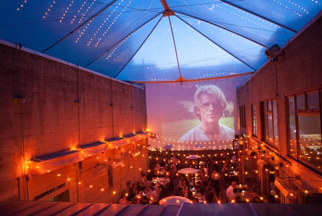 San Francisco Restaurants rooftop dining area with string lights, tables, and an outdoor movie screen under a covered evening patio.