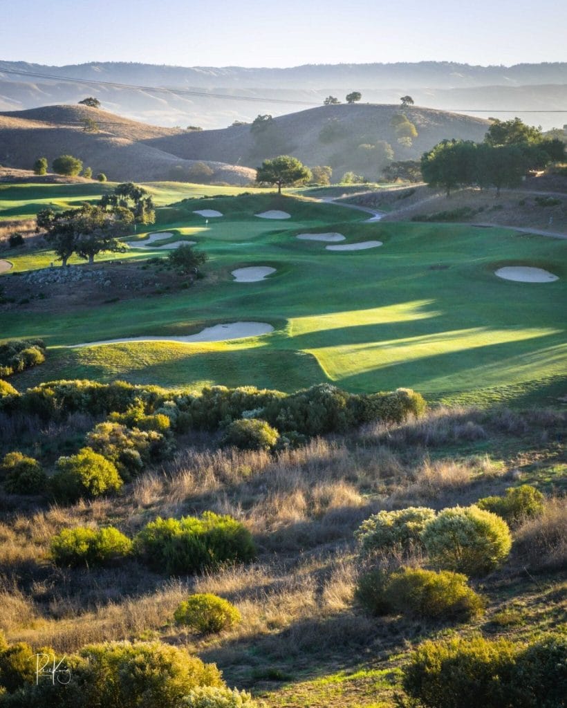 Golf course with rolling fairways, sand bunkers, shrubs, and hills in the background under soft sunlight.