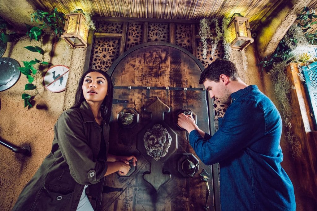 Two people working together to solve clues in a themed escape room with a large wooden door and wall props.