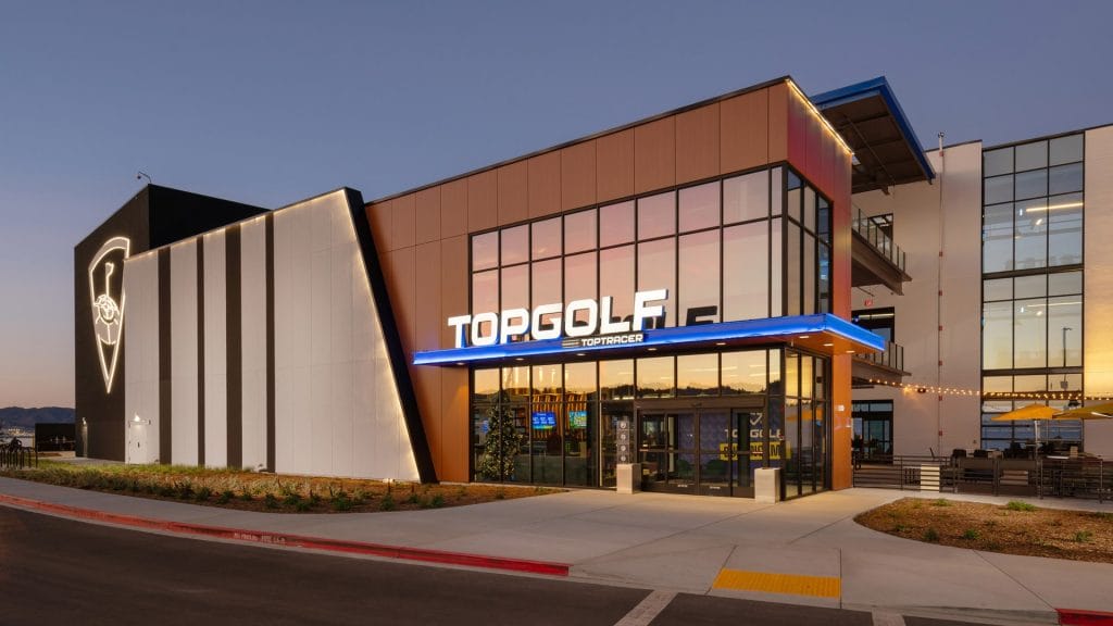 Exterior of a Topgolf venue at dusk with the illuminated Topgolf sign above the entrance and outdoor seating visible on the right.
