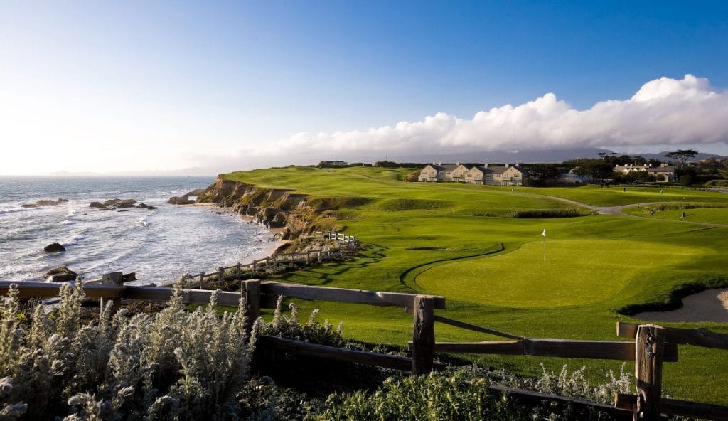 Coastal golf course with green fairways, ocean views, cliffs, and a flag on the putting green under a partly cloudy sky.