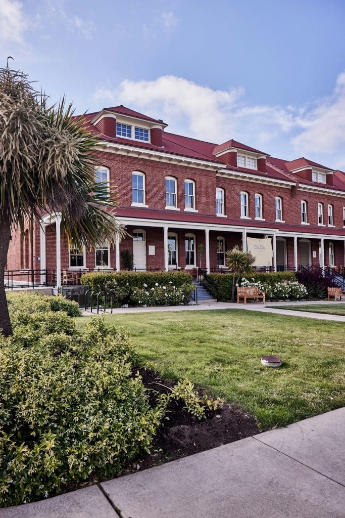 San Francisco Restaurants exterior with a red brick building, outdoor seating, greenery, and a landscaped walkway under a clear sky.