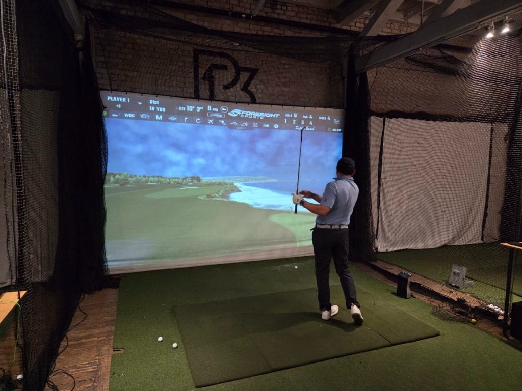 Golfer standing in an indoor simulator bay after a swing, with a virtual coastal golf course displayed on a large projection screen.