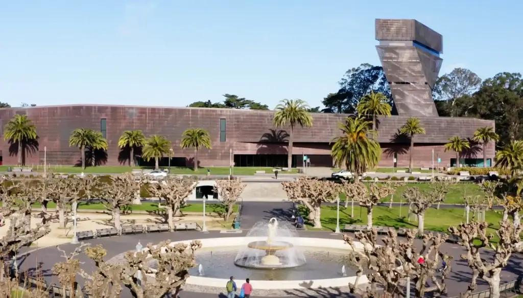 Exterior of the de Young Museum in Golden Gate Park, with palm trees, a fountain, and the museum tower in the background.