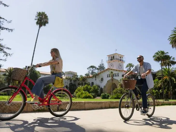 two people riding bicycles on a sunny day past a colonial style building with white clock tower, while exploring Santa Barbara