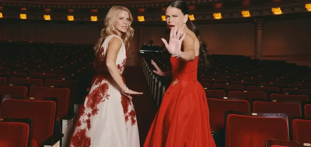 Two performers in formal dresses posing inside a concert hall with rows of red theater seats behind them.