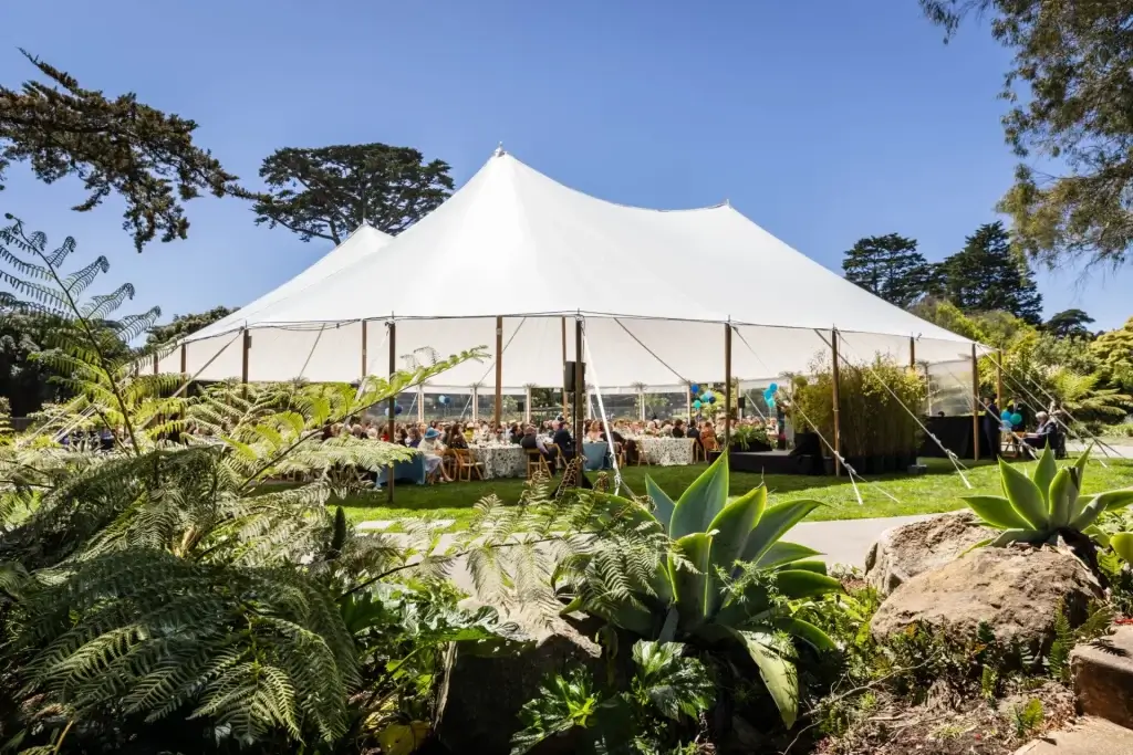 Large white event tent set up in a garden surrounded by greenery, trees, and seated guests.