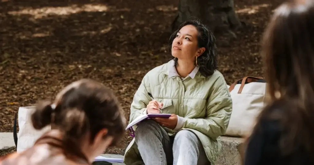 Person writing in a notebook during an outdoor poetry workshop in a wooded park setting.