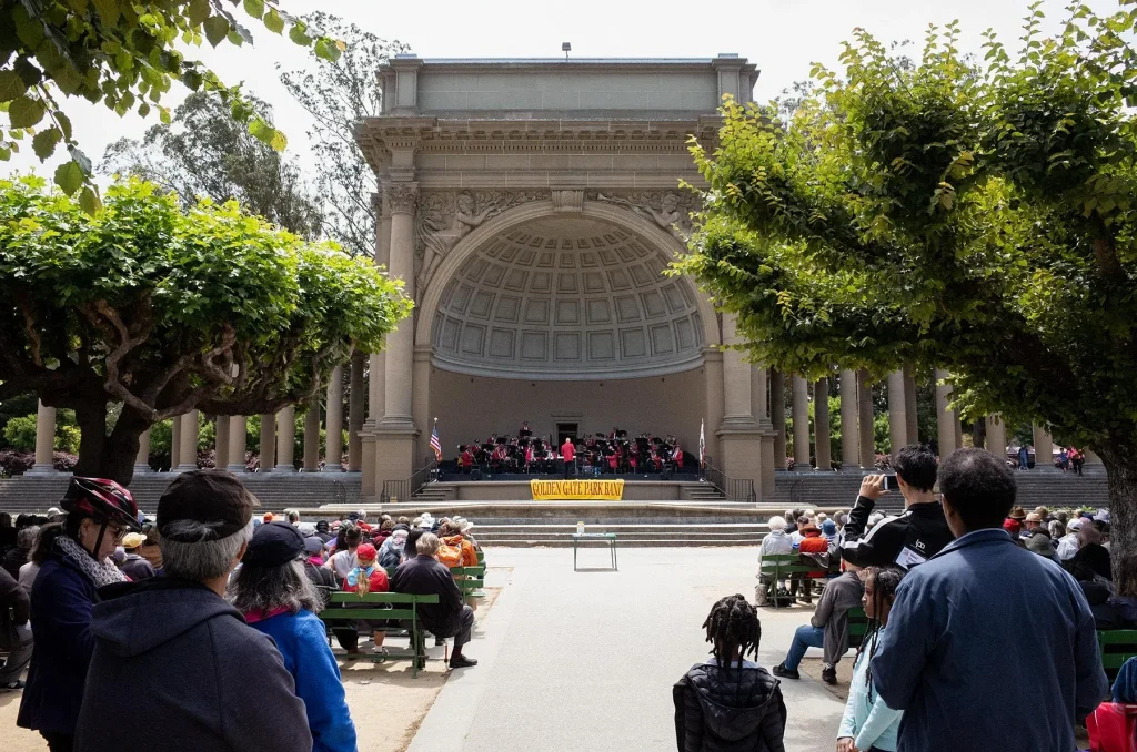 Golden Gate Park Band performing at the Spreckels Temple of Music in San Francisco, with an audience seated outdoors in Golden Gate Park.