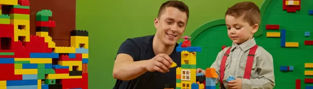 Adult and child building colourful LEGO DUPLO structures together in a play area with green walls.

